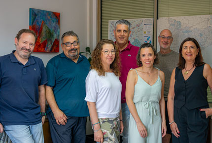 Grupo de siete personas posando juntas frente a un mapa y un cuadro abstracto en una oficina